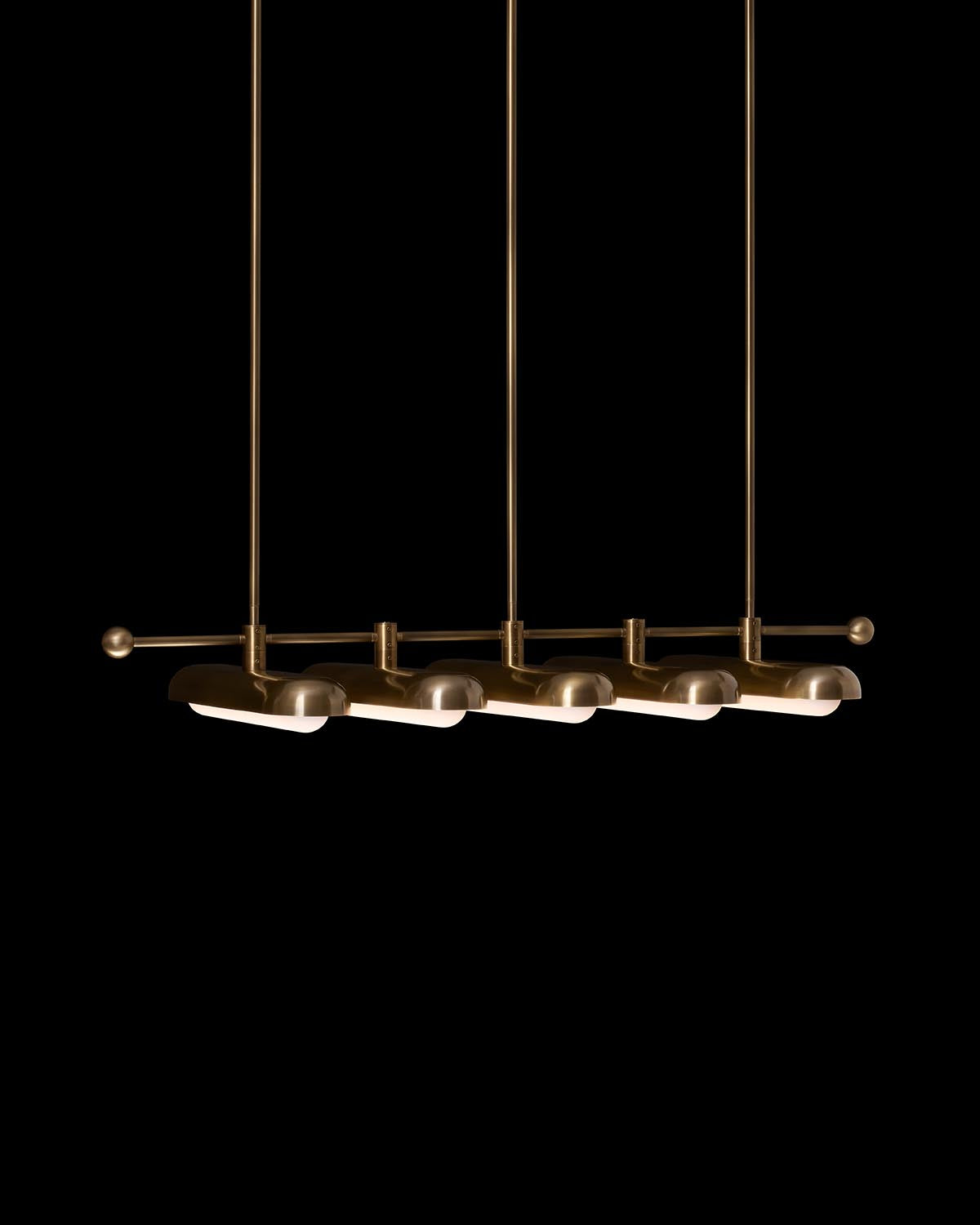 Minimalist linear pendant light with five cylindrical brass fixtures suspended by rods against a black background