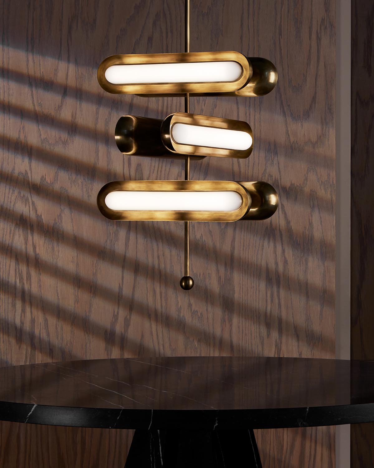 Three horizontal brass tube lights with frosted glass shades mounted on a dark wood wall above a black marble table