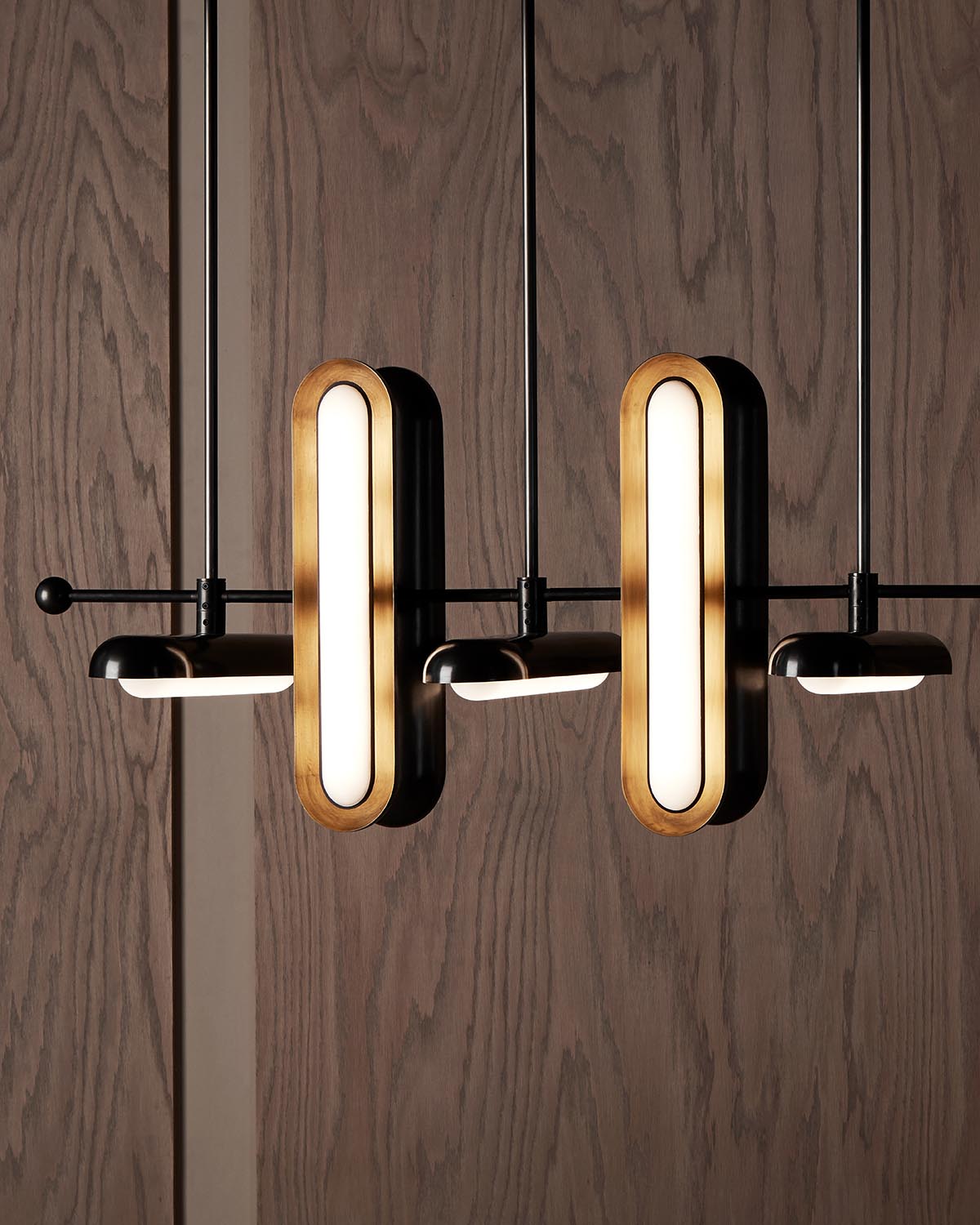 Close-up of modern pendant light fixtures with elongated oval wood frames and black metal arms against a dark wood wall
