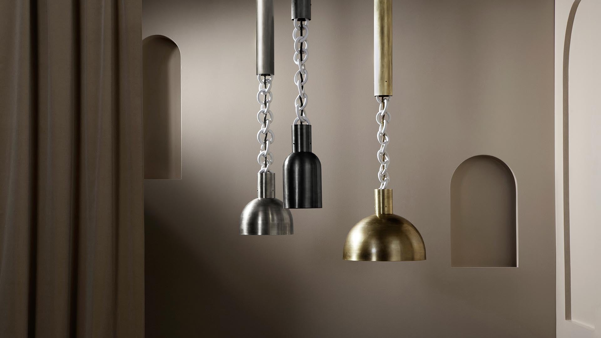 Three pendant lights with metallic finishes hanging by chain links in front of a taupe wall with arched recesses