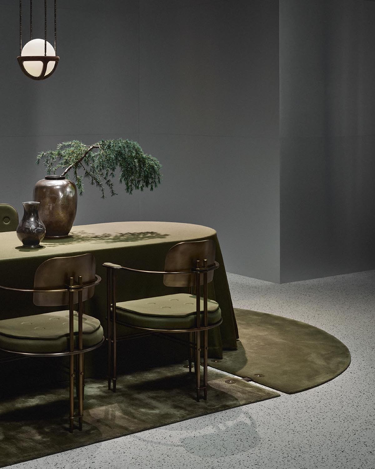 Minimalist interior with oval olive-green table covered in fabric, two bronze chairs, green rug, and modern pendant light
