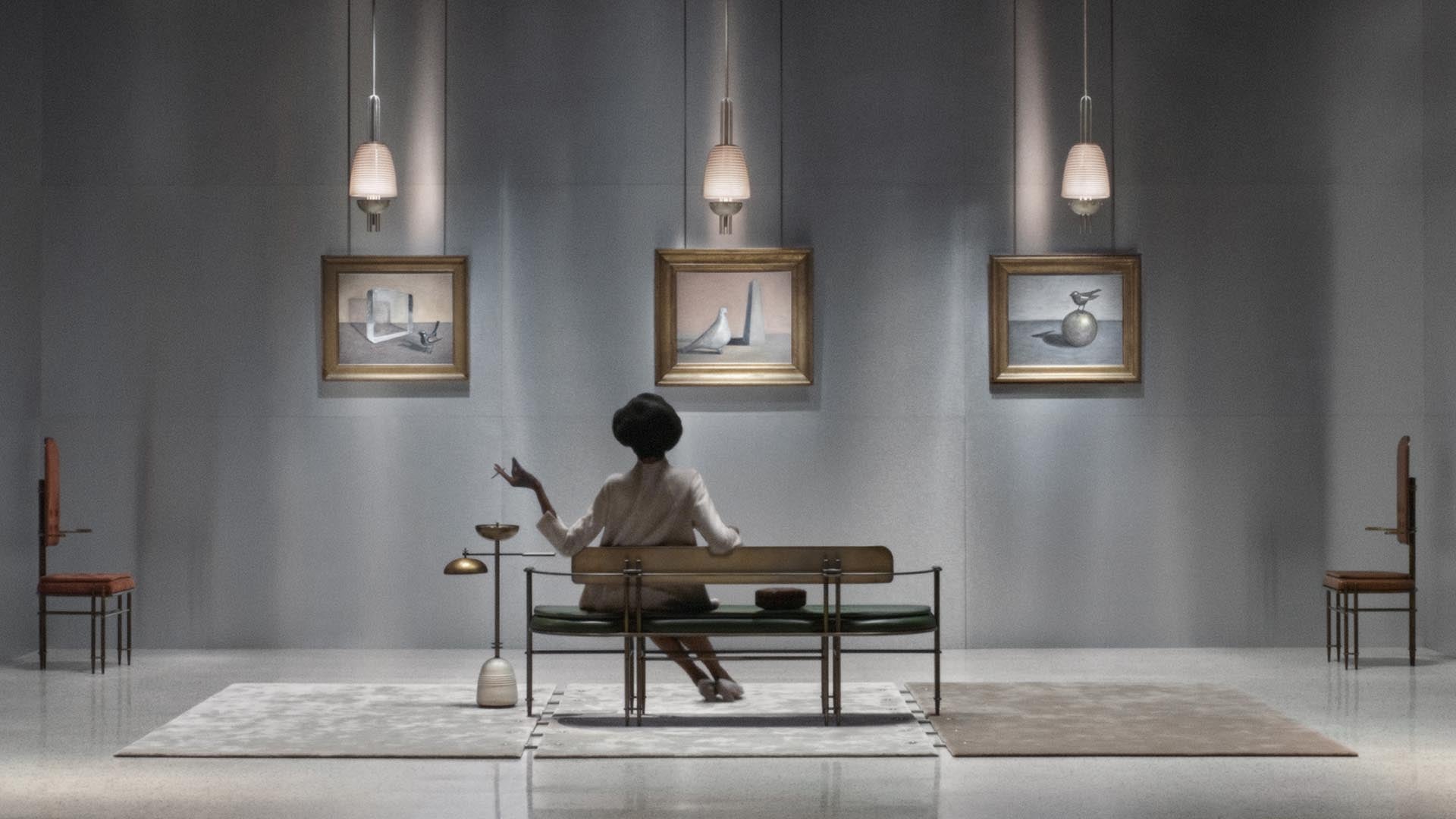 Woman seated on a bench in a minimal gallery space with three framed paintings and pendant lights above