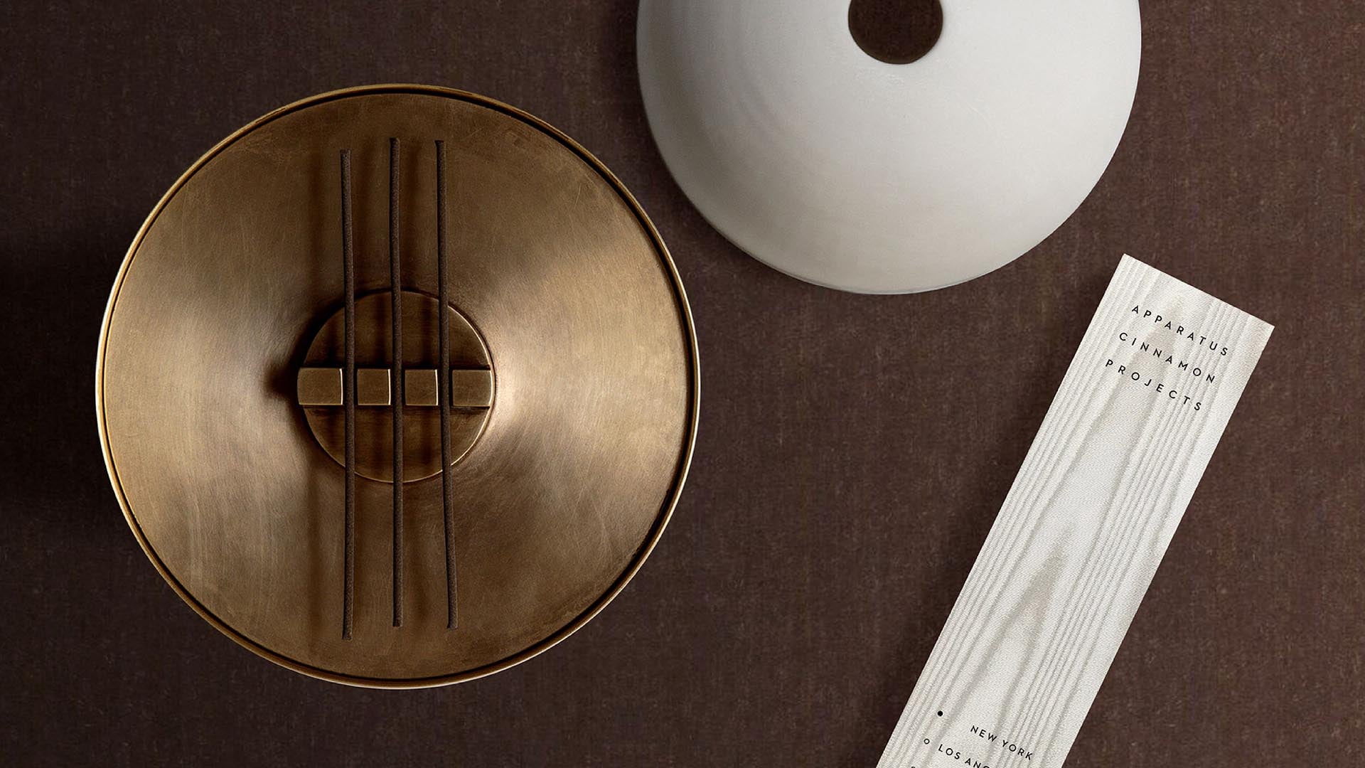 Overhead view of a bronze circular sculpture with vertical rods, a white round ceramic vase, and a textured paper strip on a dark surface