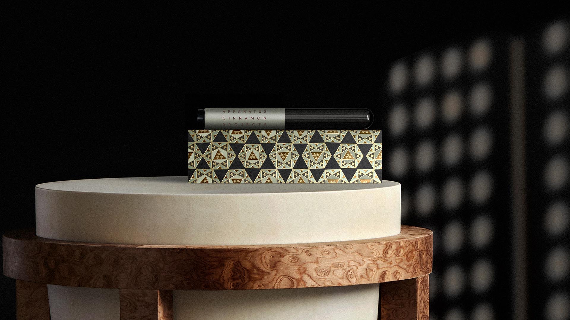 Rectangular box with geometric pattern and black pen placed on round white pedestal with wooden base against dark background