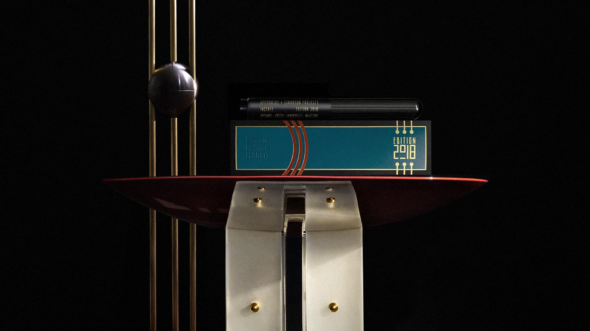 Close-up of a dark teal rectangular tin with gold lettering and a black pen resting on top, displayed on a red curved surface against black background