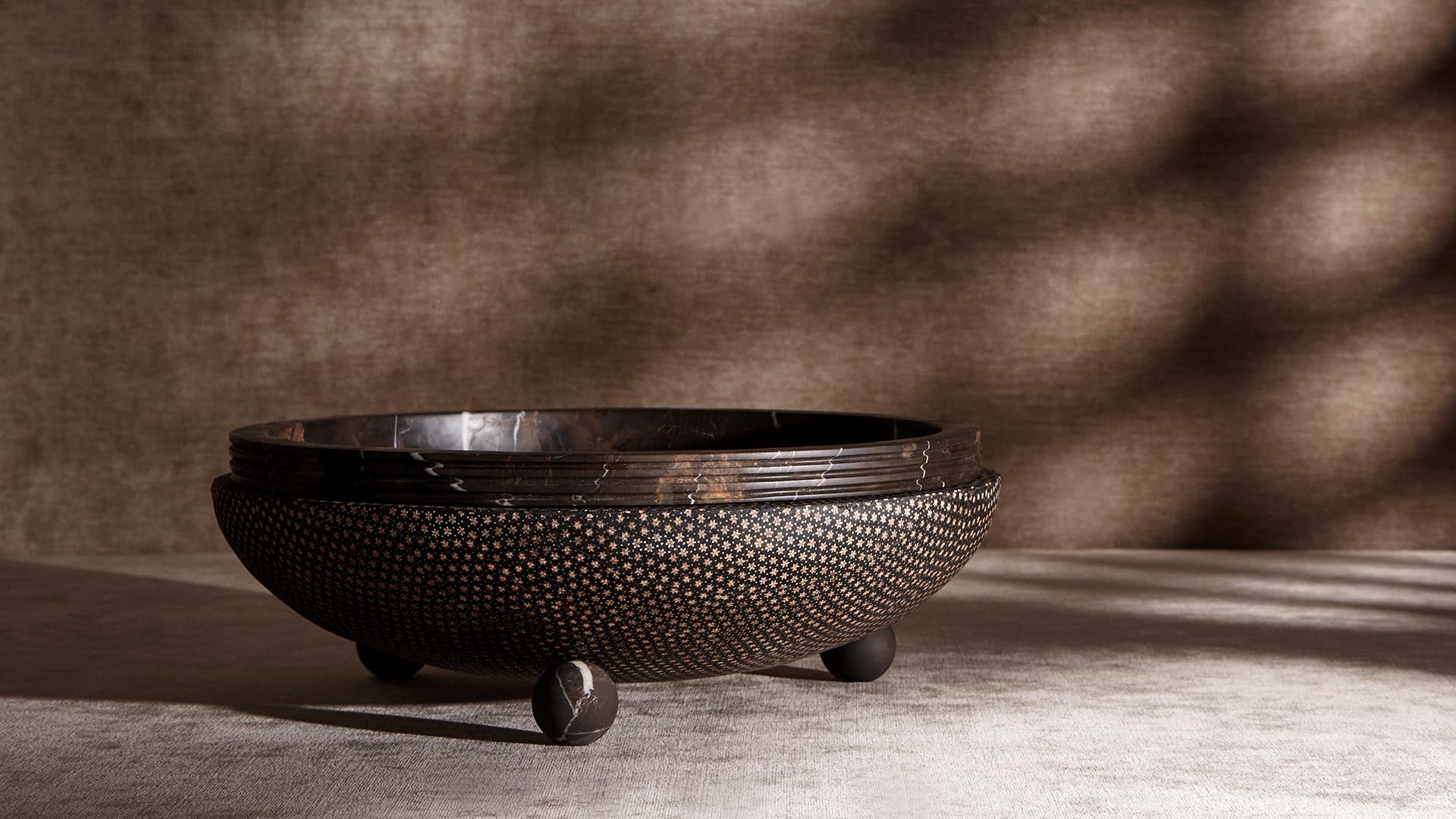 Low round decorative bowl with textured dark surface and three spherical legs casting soft shadows on a light fabric background