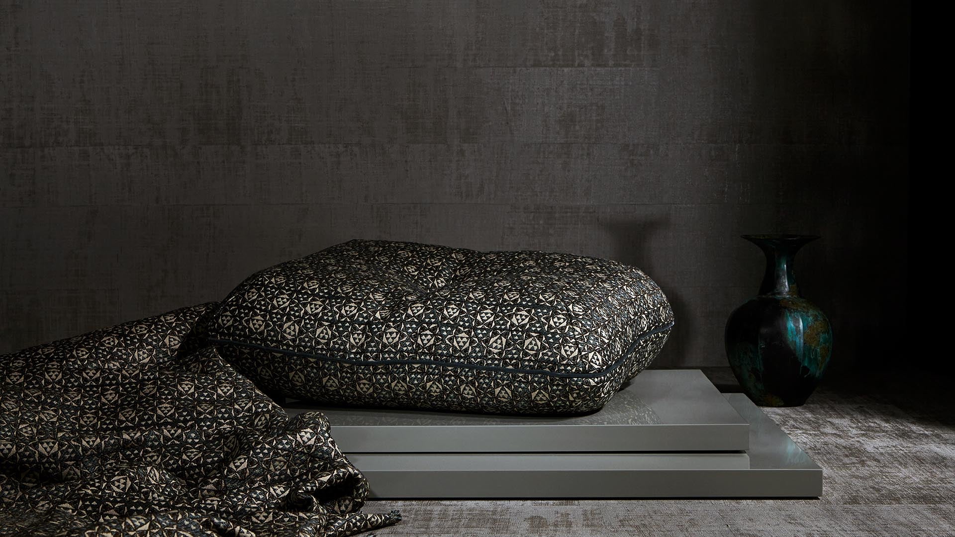 Decorative black and gold patterned pillow resting on stacked gray platforms next to a dark green ceramic vase on textured floor