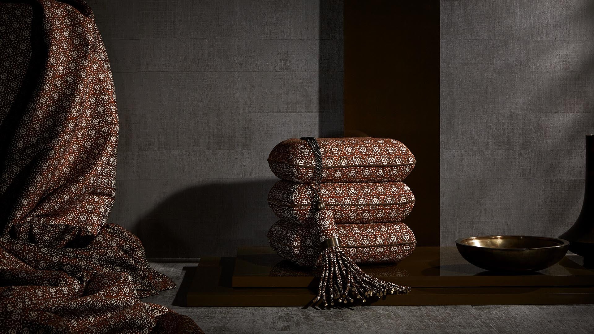 Stack of folded brown patterned fabric with tassel detail alongside matching draped fabric and brass bowl on textured surface
