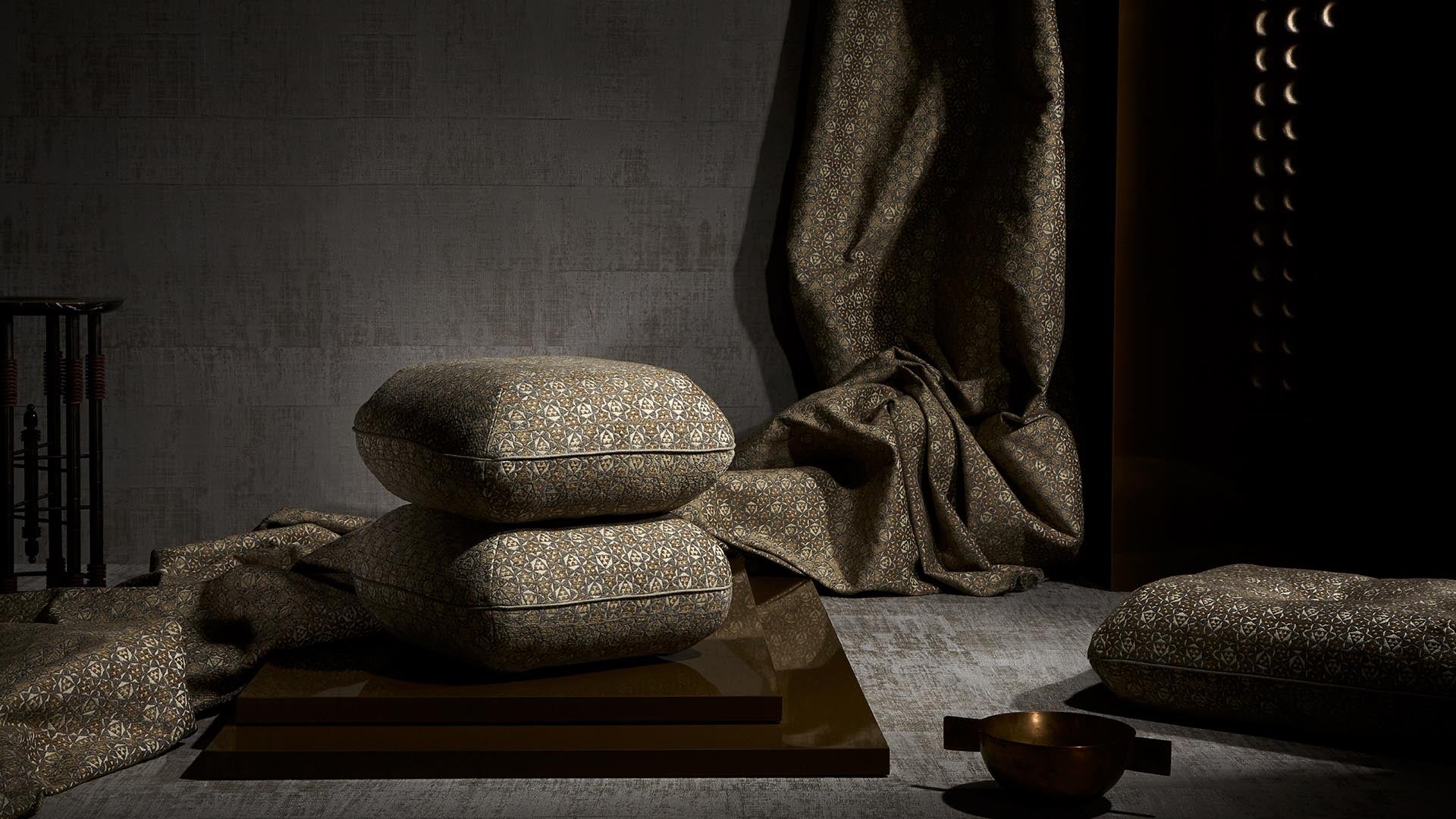 Two patterned cushions stacked on a reflective surface with matching draped fabric and a small bowl on a textured floor