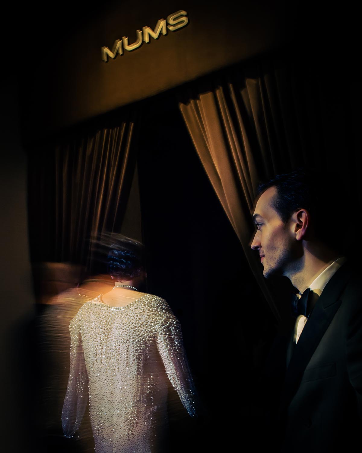 Man in tuxedo watching a woman in a pearl-studded dress walking through dark curtains beneath a sign reading MUMS