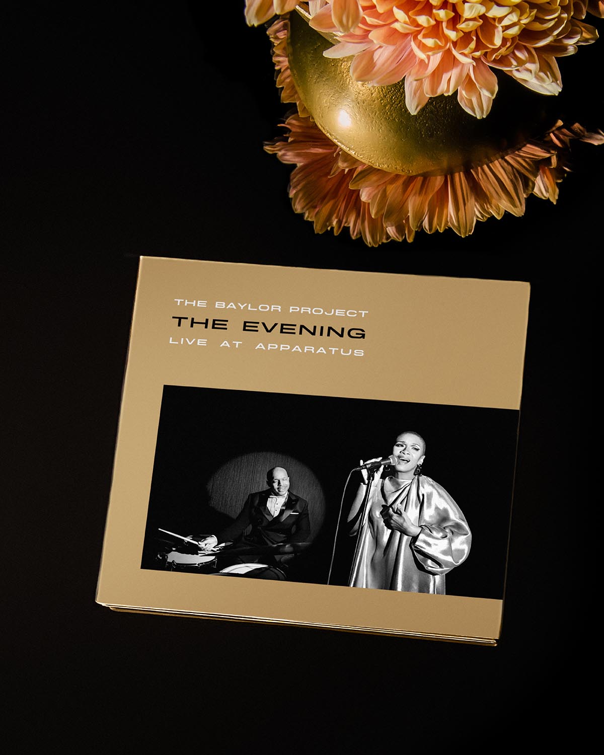 Square album cover with gold background featuring black and white photo of a singer and drummer, next to a peach flower arrangement