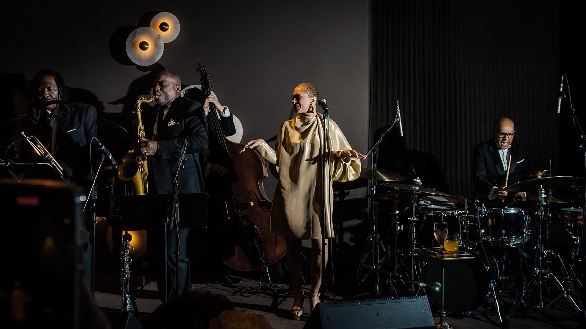 Jazz band with trumpet, saxophone, upright bass, vocalist in beige dress, and drummer performing on dimly lit stage