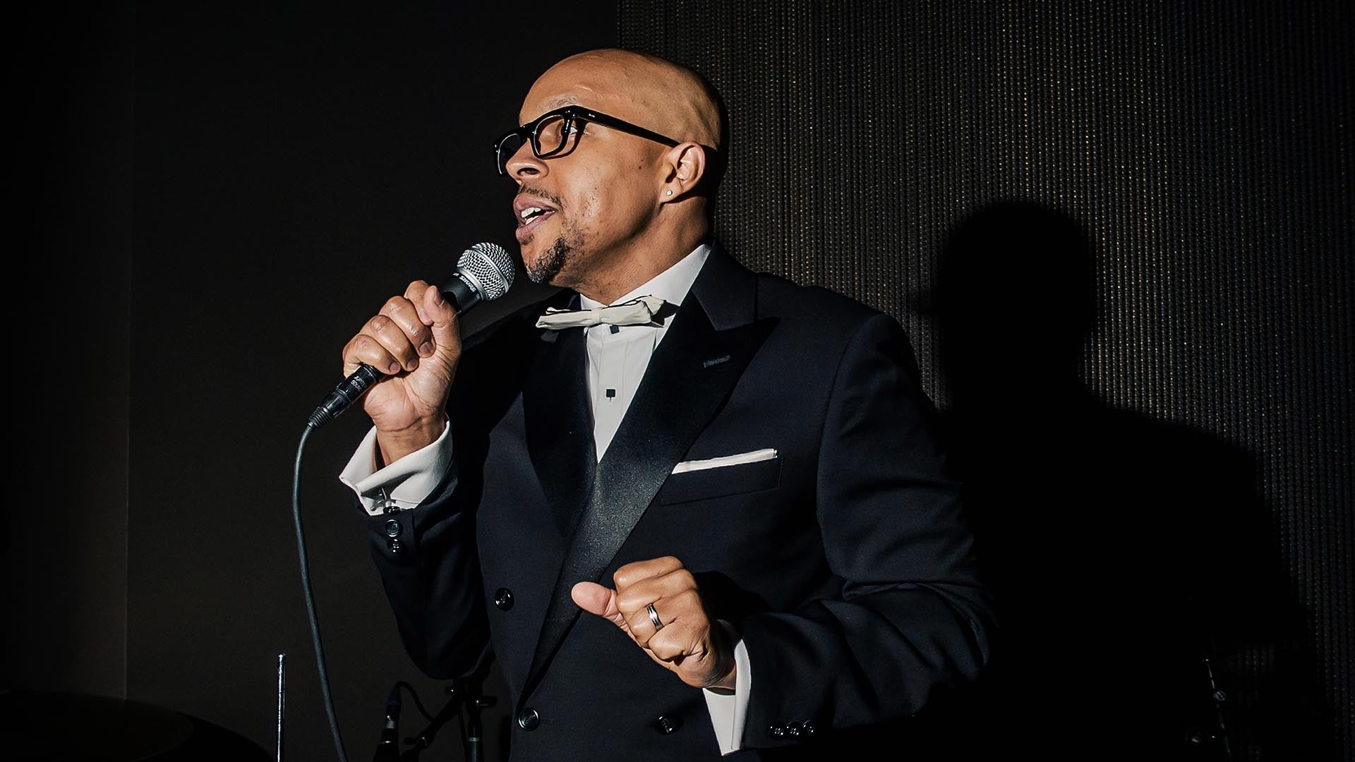 Man in black tuxedo and glasses singing into a microphone against a dark background with shadow behind him
