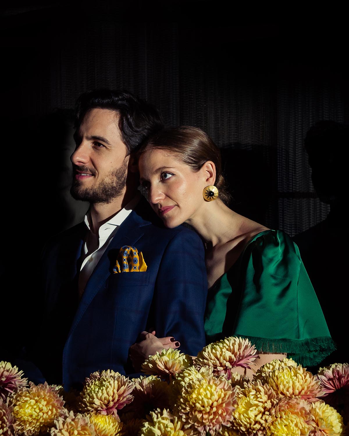 Man in blue suit with yellow pocket square and woman in green dress leaning on his shoulder surrounded by flowers