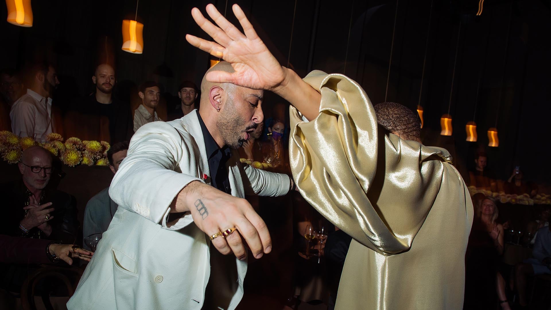 Two people dancing closely in a dimly lit room with hanging pendant lights and an audience in the background