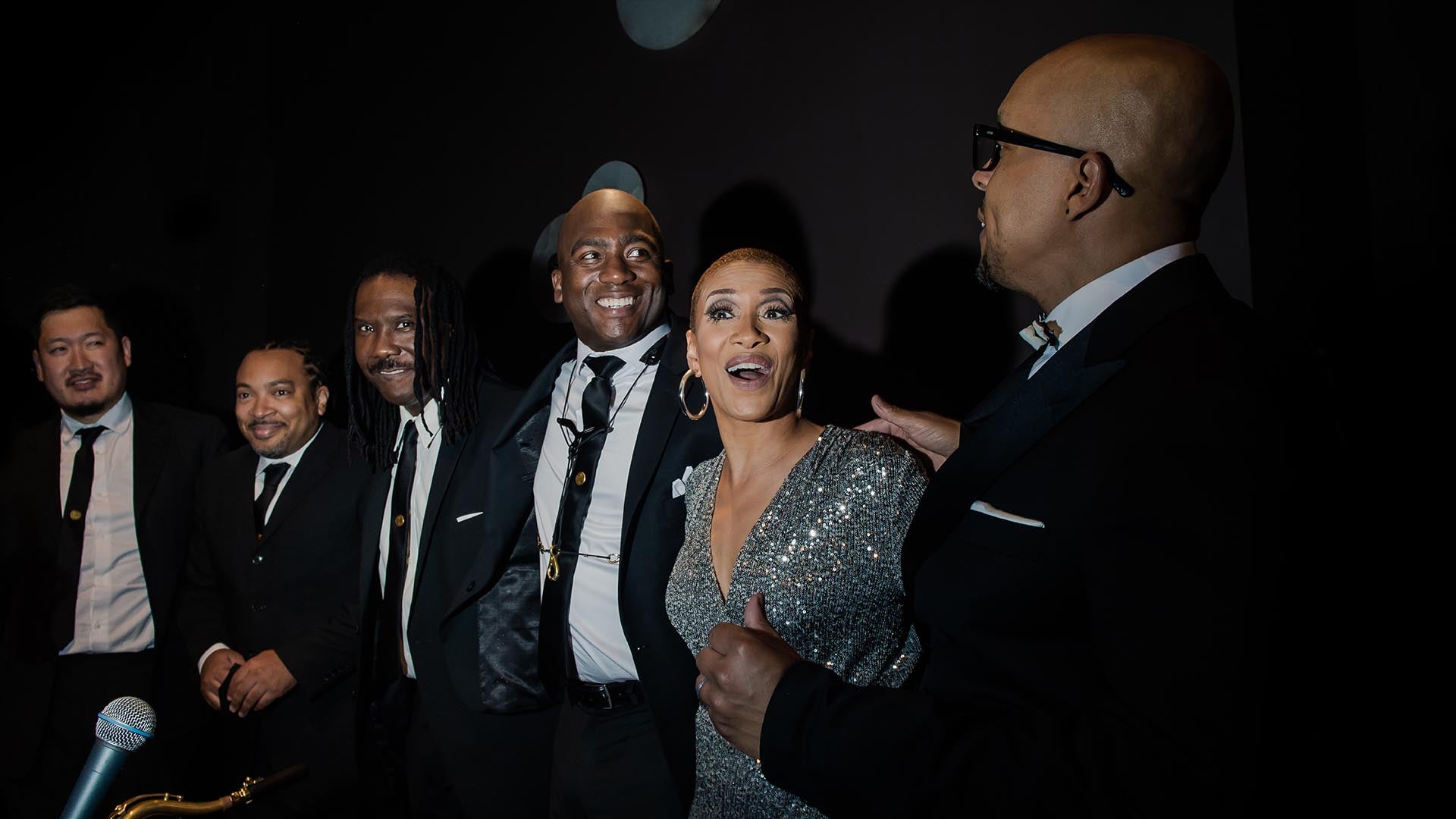 Group of six well-dressed people standing in a dark room, smiling and interacting with each other.