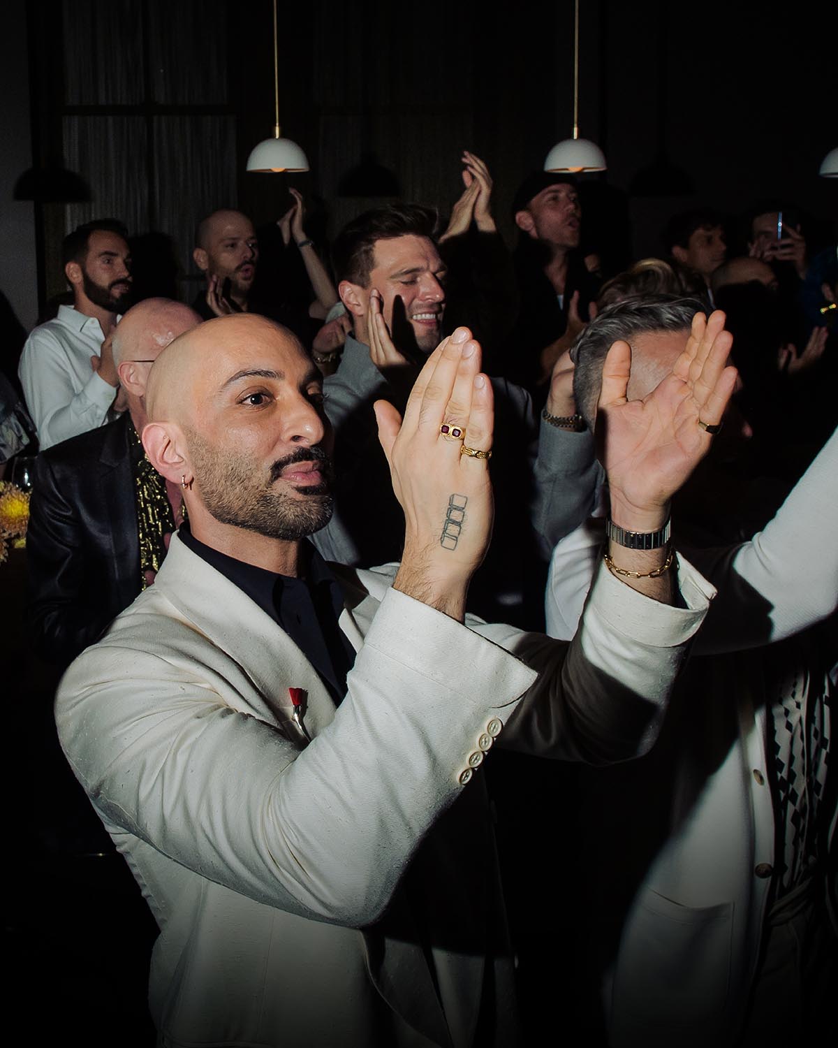 Man in light beige blazer clapping with crowd of people applauding in dimly lit indoor setting