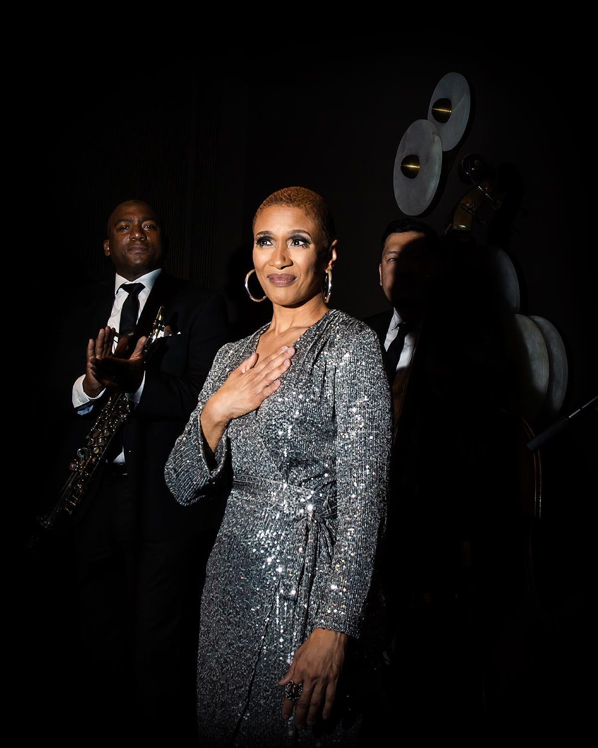 Woman in a glittering silver dress with hand on chest, standing in spotlight with two men in dark suits behind her
