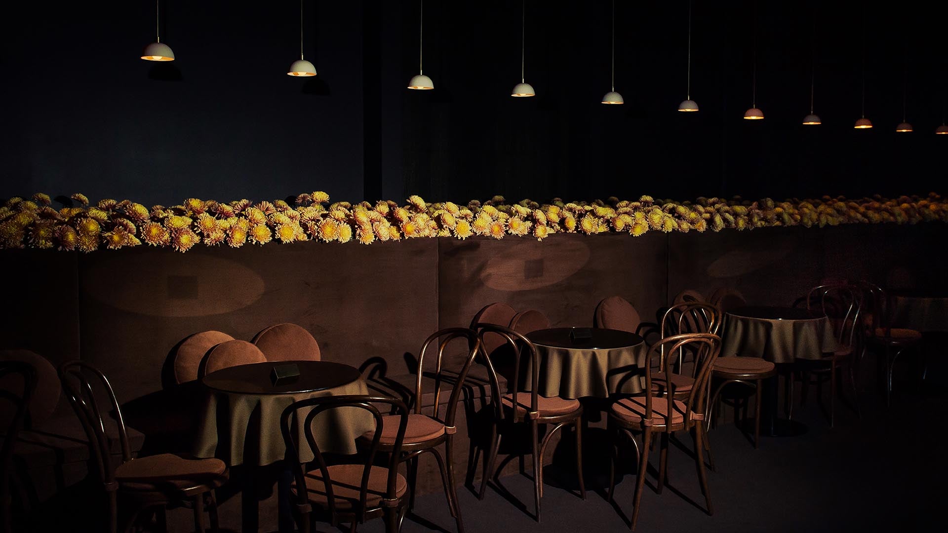Dimly lit café interior with wooden chairs and tables draped in dark cloth, accented by a wall lined with yellow flowers