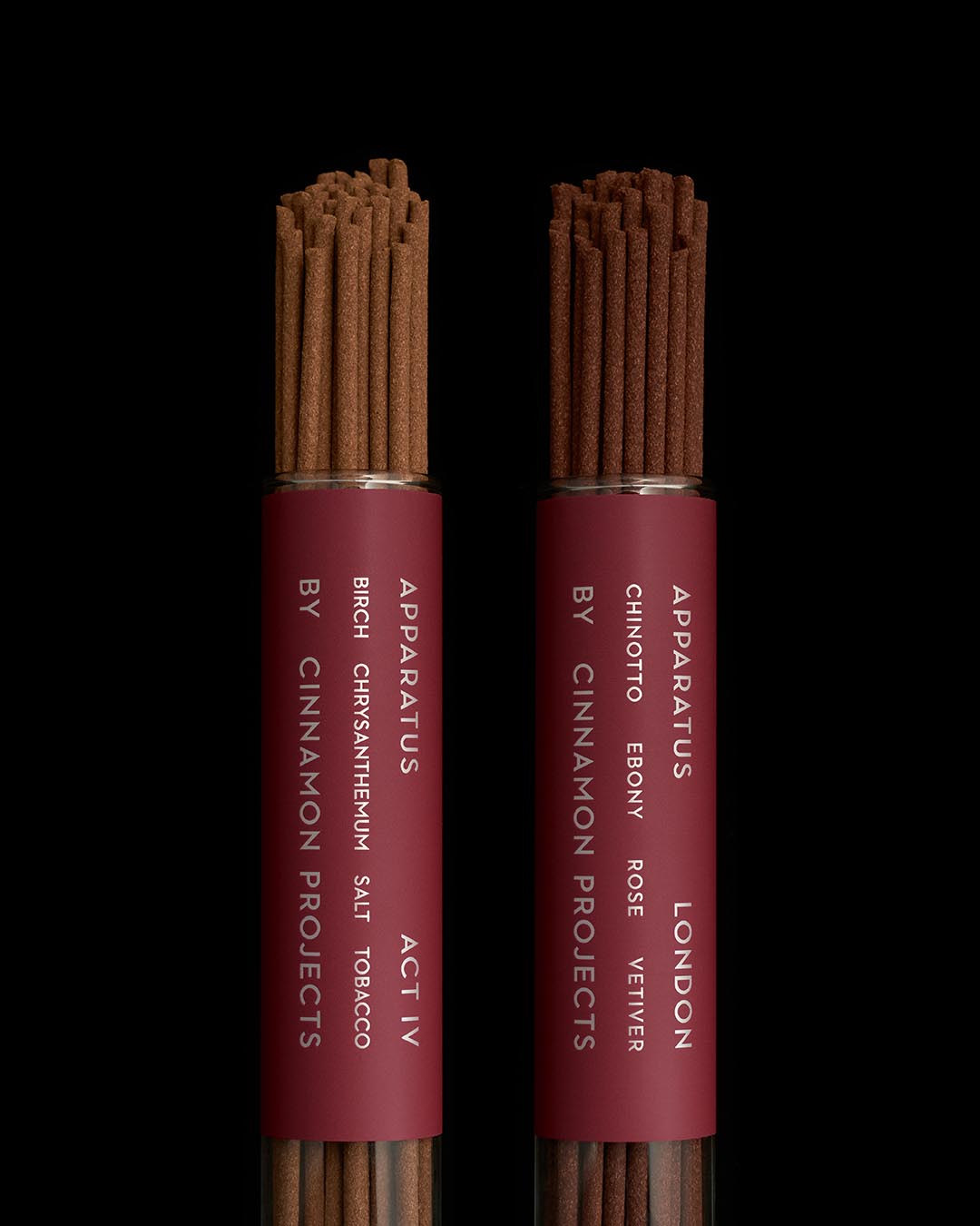 Two cylindrical burgundy tubes filled with brown incense sticks labeled Apparatus by Cinnamon Projects against a black background