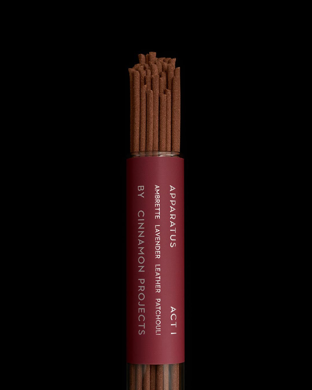 Close-up of a cylindrical glass tube filled with brown incense sticks labeled Apparatus Act I on a black background