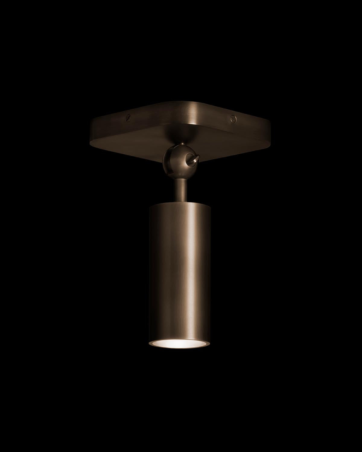 Close-up of a cylindrical downlight fixture with matte bronze finish mounted on a square ceiling plate against black background
