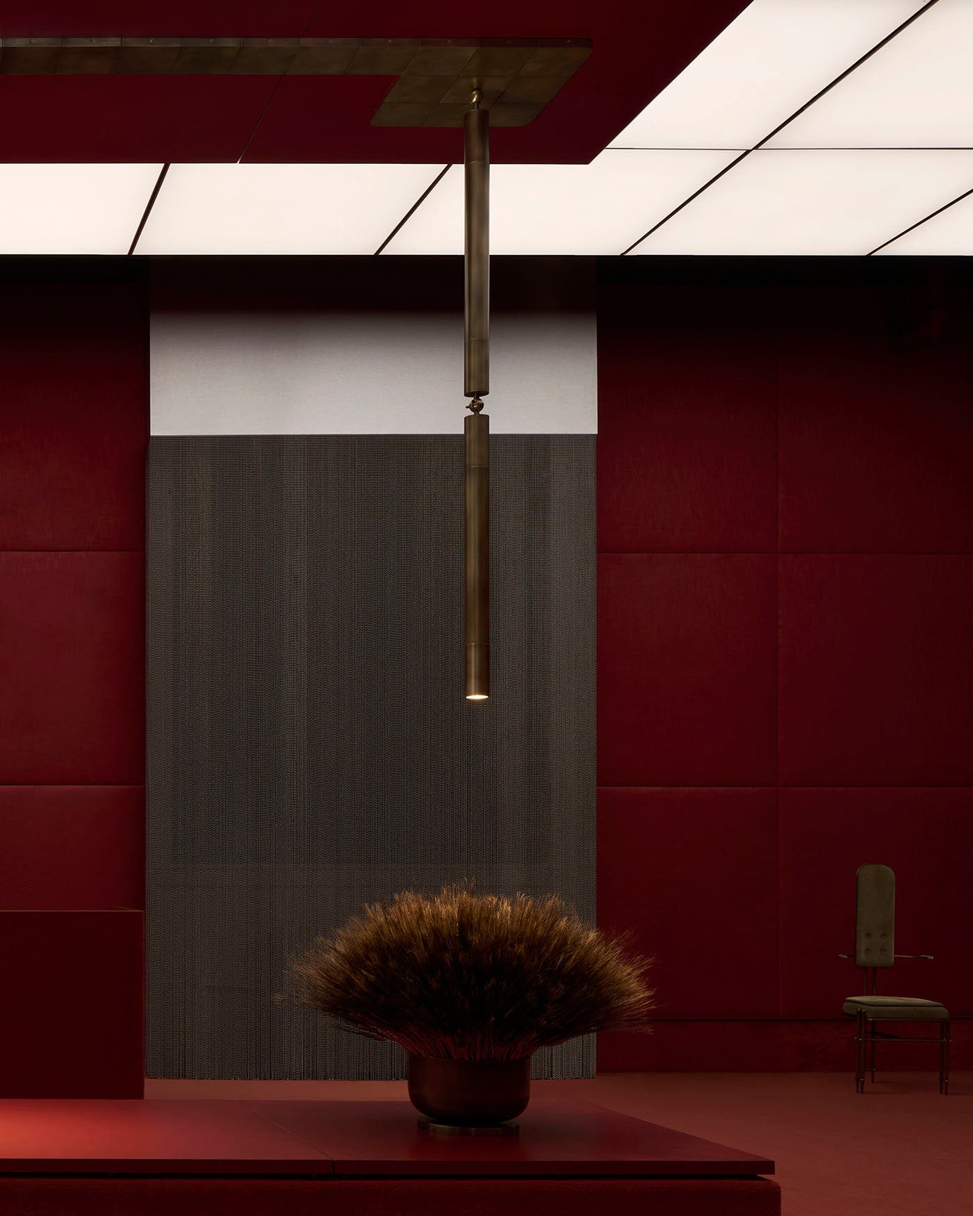 Slim brass cylinder pendant light hanging from a ceiling above a low table with dried floral arrangement in a red and dark wood room