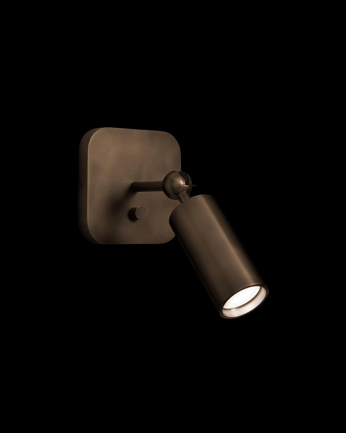Close-up of a bronze cylindrical wall sconce light with adjustable head, mounted on a square base against black background