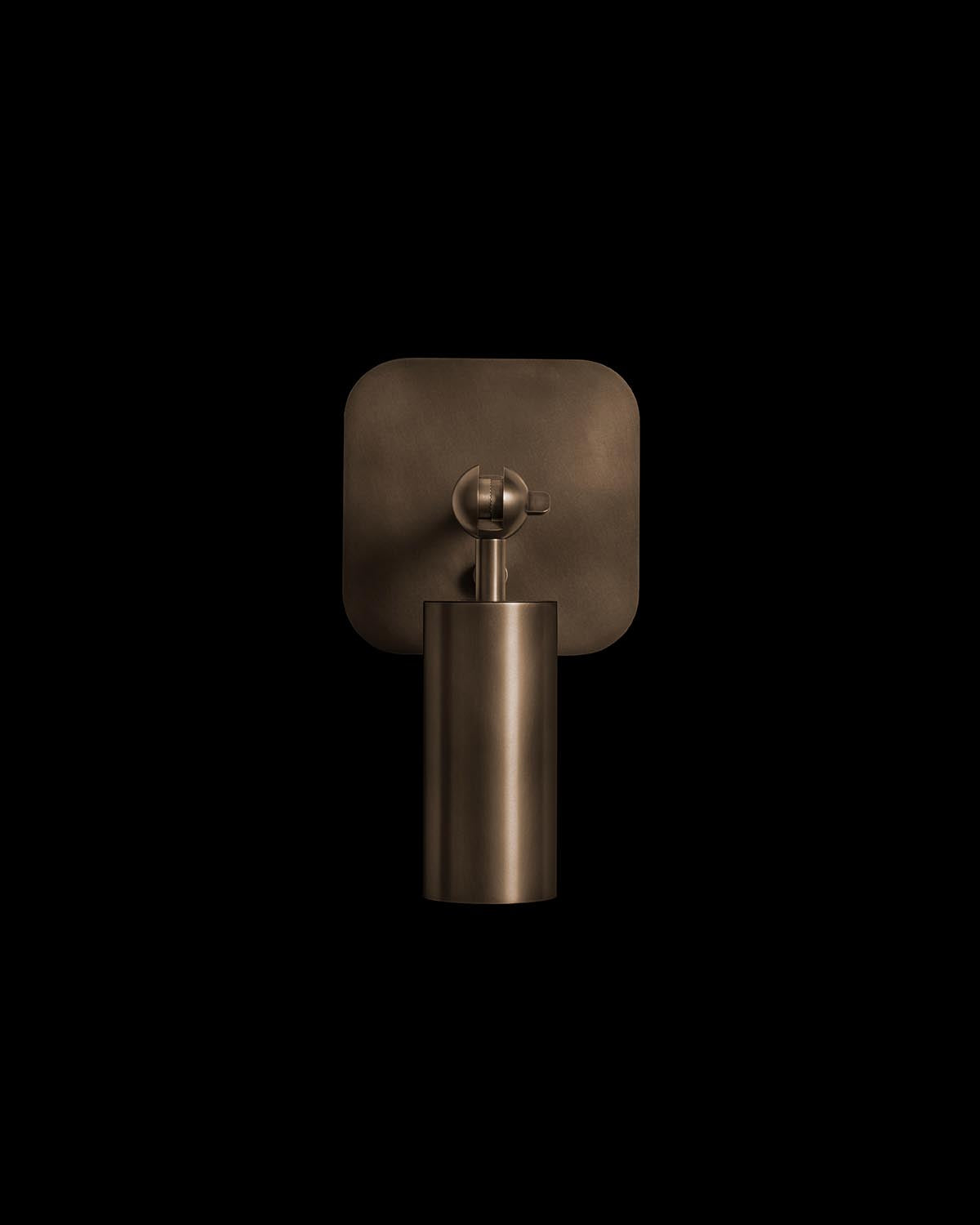 Minimalist bronze cylinder wall sconce with rounded square backplate against a black background