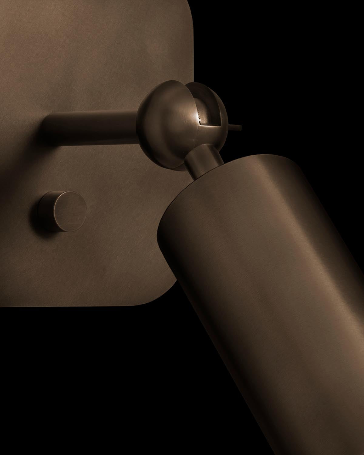 Close-up of a matte bronze cylindrical wall sconce with adjustable joint mounted on a square backplate against black background