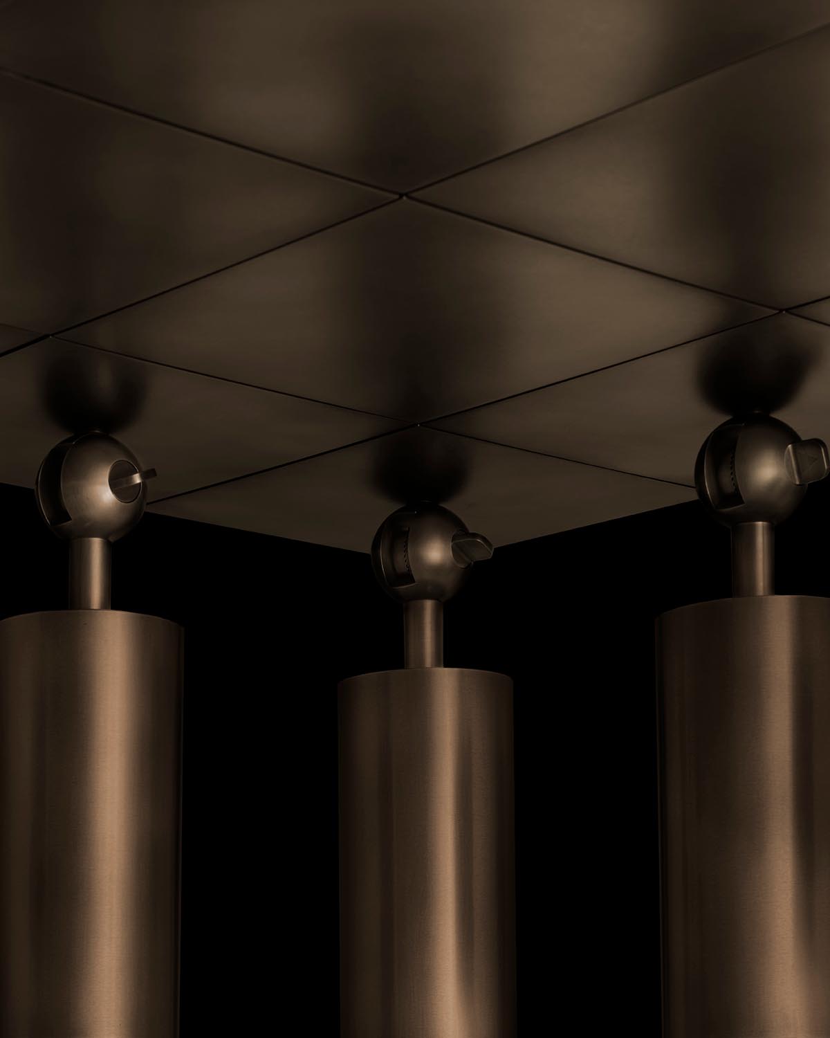 Close-up of three vertical bronze cylinder fixtures with round tops mounted under a geometric metal ceiling panel