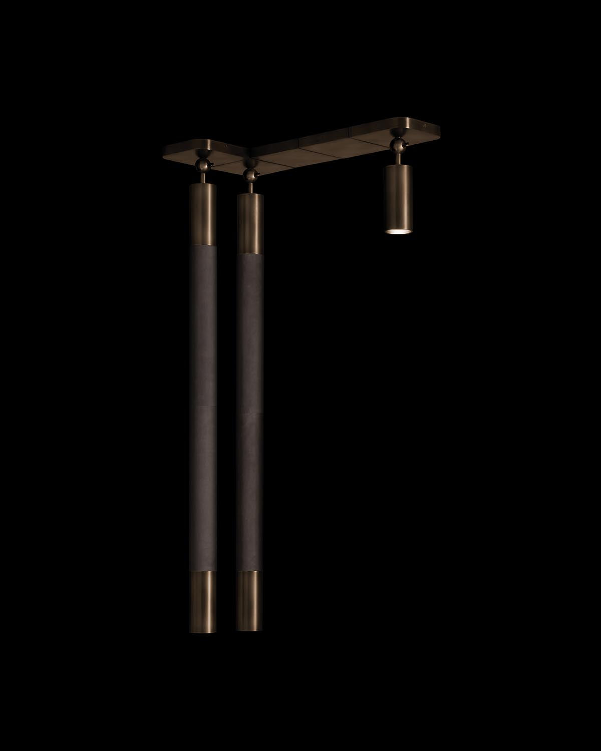 Minimalist ceiling light fixture with two elongated cylindrical pendants and one smaller spotlight in brass finish on black background