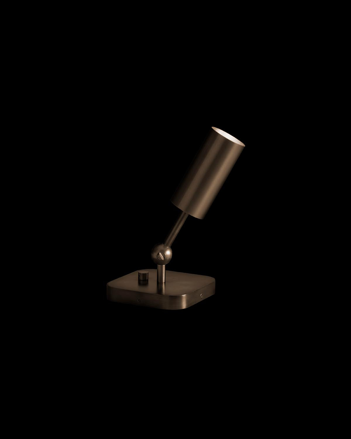 Minimalist bronze cylindrical uplight with adjustable head on square base against a black background