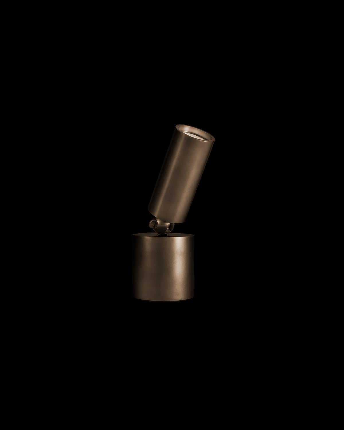 Minimalist bronze cylindrical uplight fixture with adjustable angled head on matching base against black background