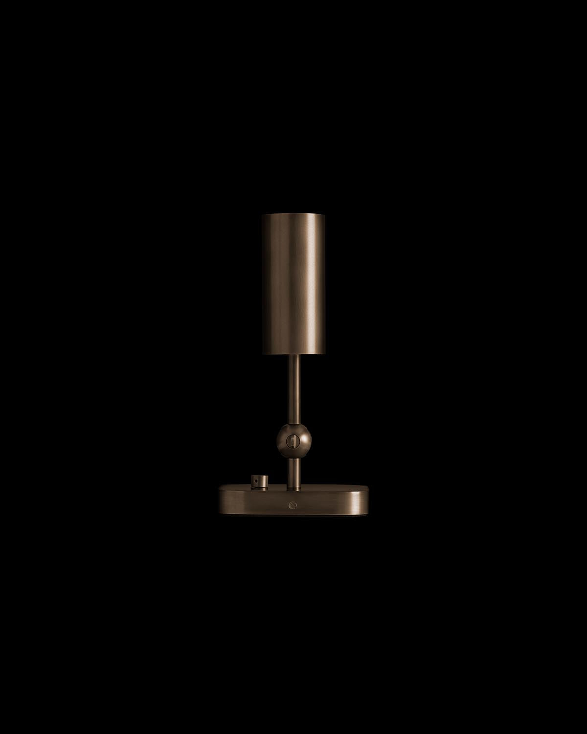 Minimalist bronze uplight lamp with cylindrical shade and spherical accent on slim stand against black background