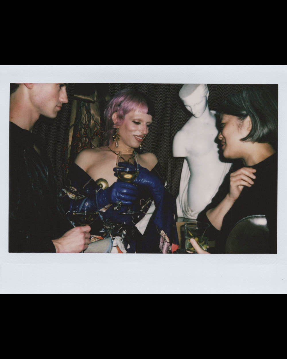 Three people engaged in conversation near a white classical statue in a dimly lit indoor setting.
