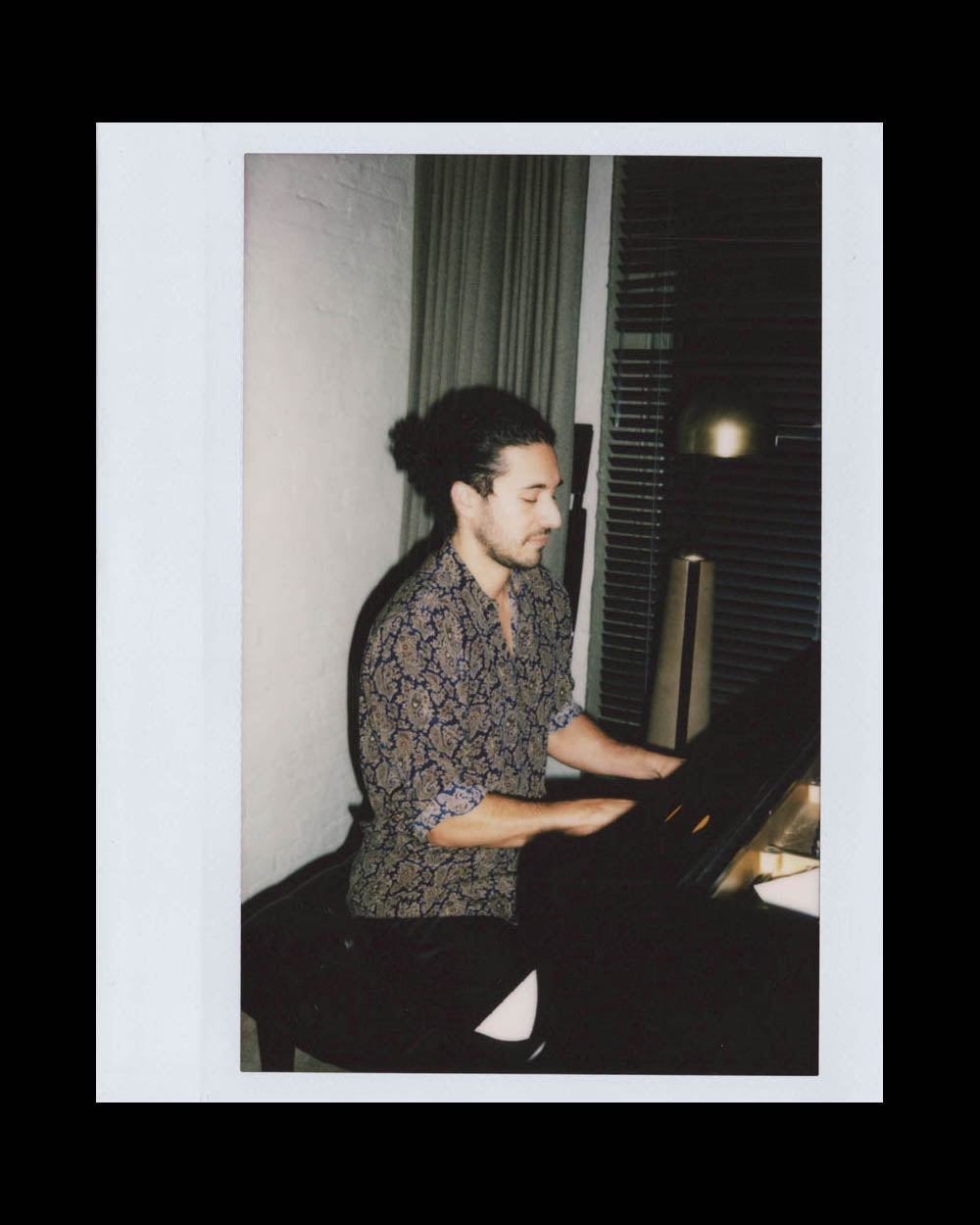 Man with tied-back hair playing piano in a dimly lit room with curtains and blinds behind him