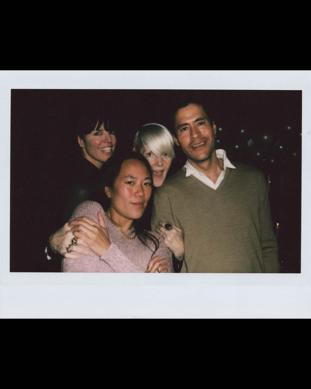 Group of four adults smiling closely together in dim indoor setting with dark background