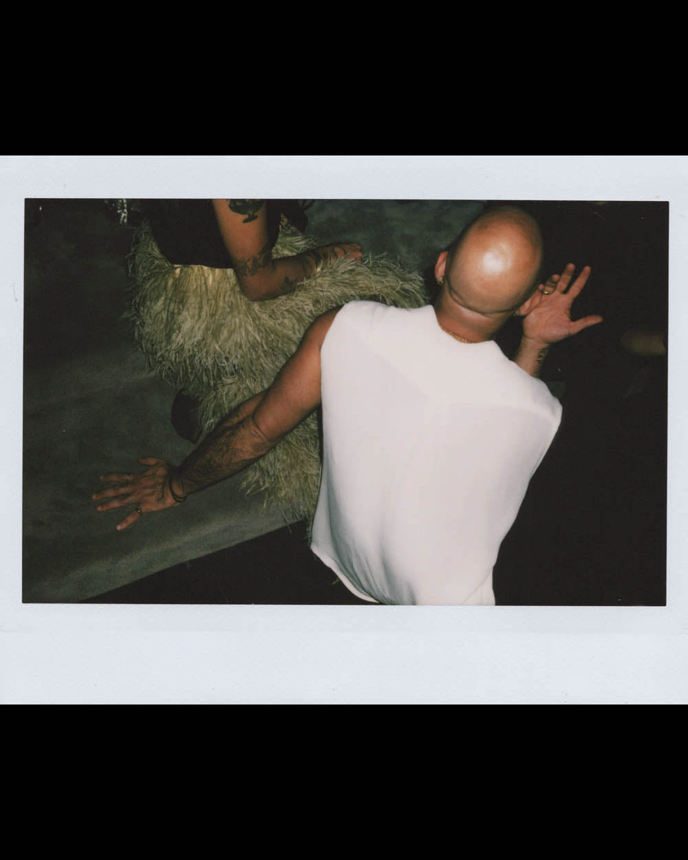 Bald man in white sleeveless shirt gesturing with hand while sitting next to person wearing textured clothing in dark setting