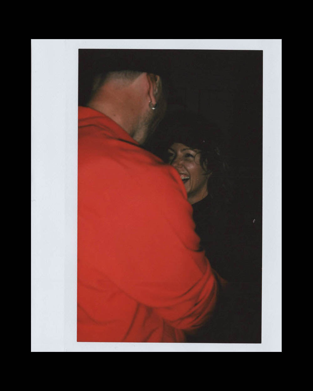 Man wearing red jacket facing a smiling woman in a dark indoor setting, sharing a joyful moment.