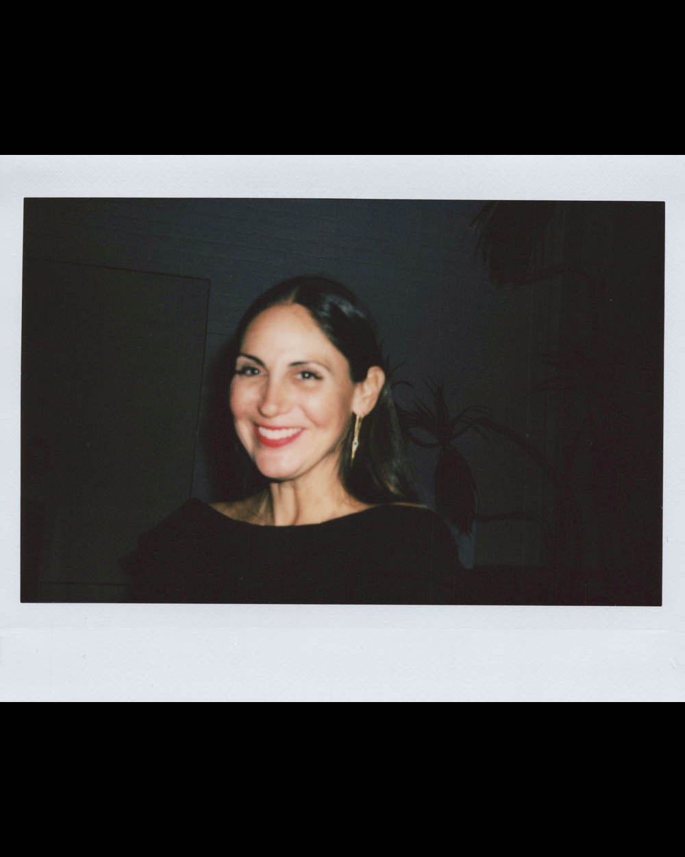 Woman with long dark hair smiling against a dark background wearing a black top and gold earrings