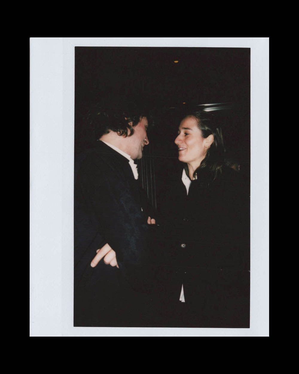 A man and a woman face each other smiling and talking in a dimly lit indoor setting.
