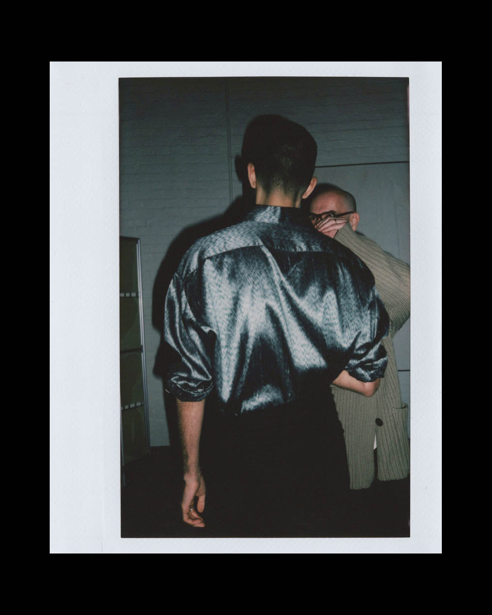 A man in a shiny dark shirt with rolled sleeves stands with his back to the camera while another man adjusts his glasses nearby.