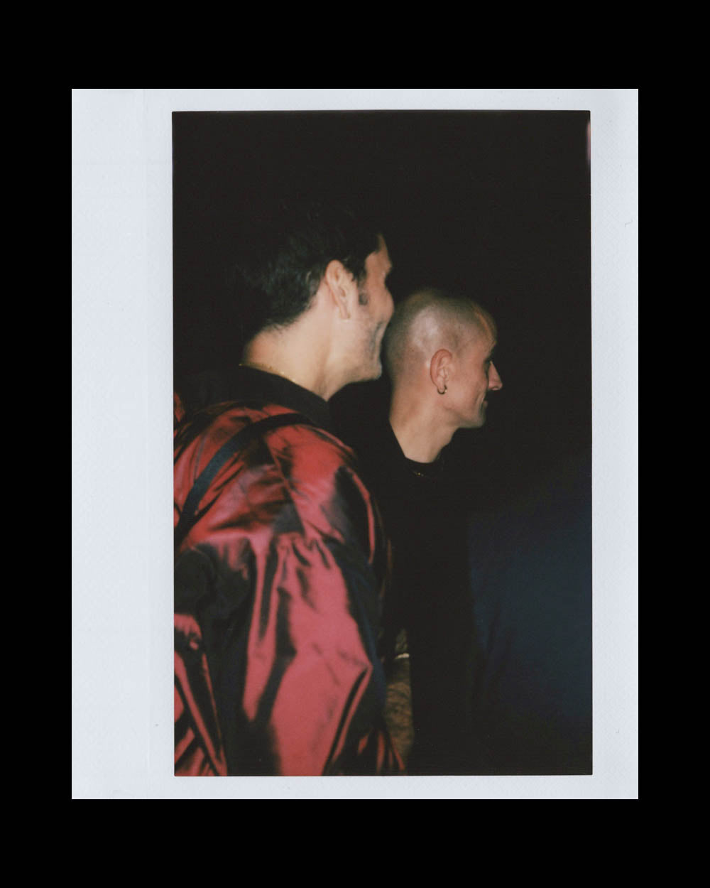 Two men facing sideways and smiling, one wearing a red satin jacket and the other with a shaved head against a dark background