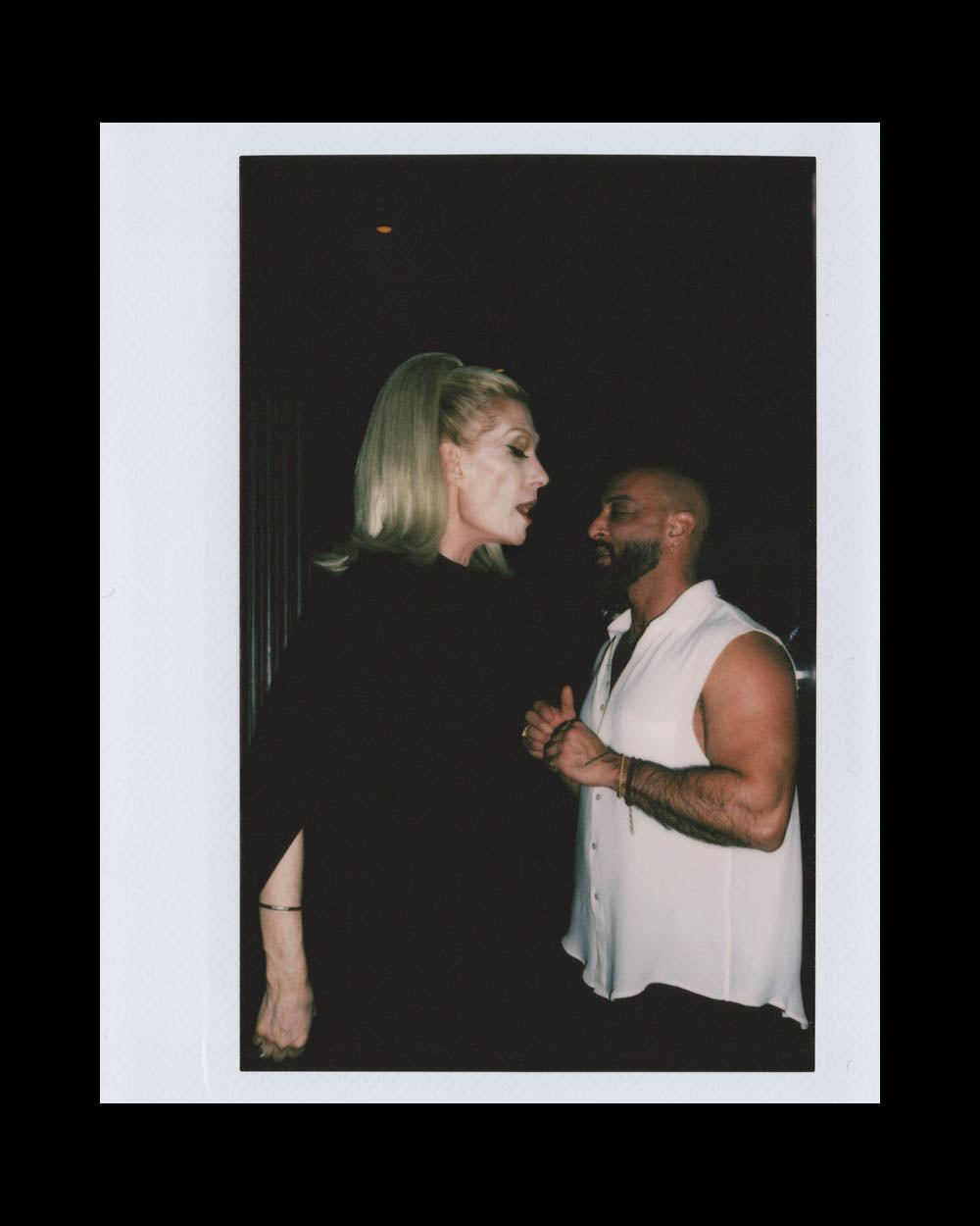 A tall woman with blonde hair talking to a muscular man in a sleeveless white shirt in a dark setting.