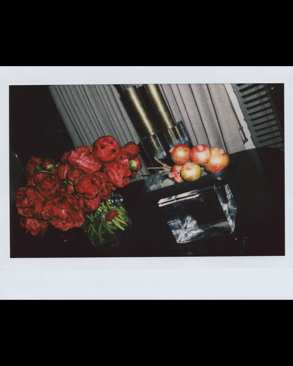 Red peonies in a glass vase next to a bowl of apples on a glossy black surface with a curtain background.