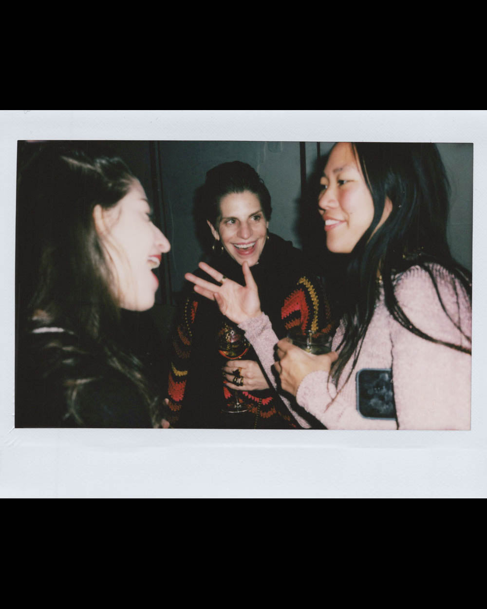 Three women smiling and talking indoors, one gesturing with her hand while holding a drink in each hand.