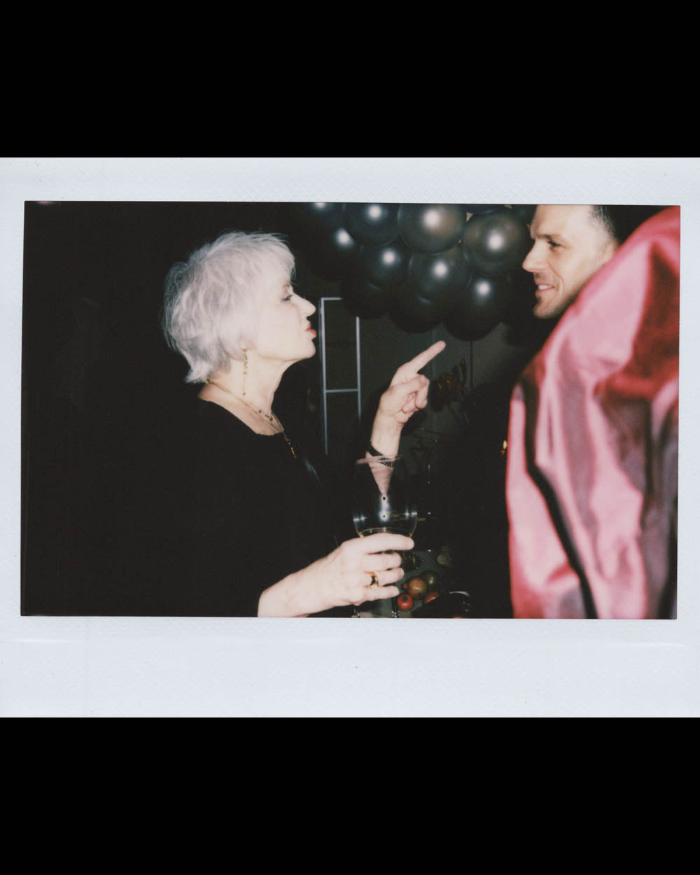 An older woman with white hair holding a glass gestures toward a smiling man in a dark room with black balloons.