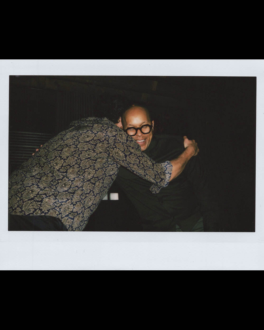 Two men warmly embracing in a dark room, one wearing glasses and smiling broadly.