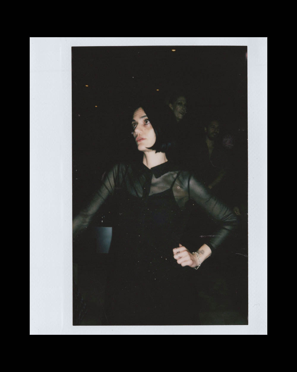 A woman with short dark hair wearing a black sheer long-sleeve top standing in a dimly lit space.